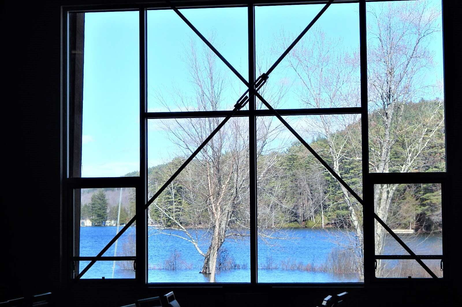 A large window frame with diagonal metal supports looks out onto a calm blue lake surrounded by trees and a hillside.