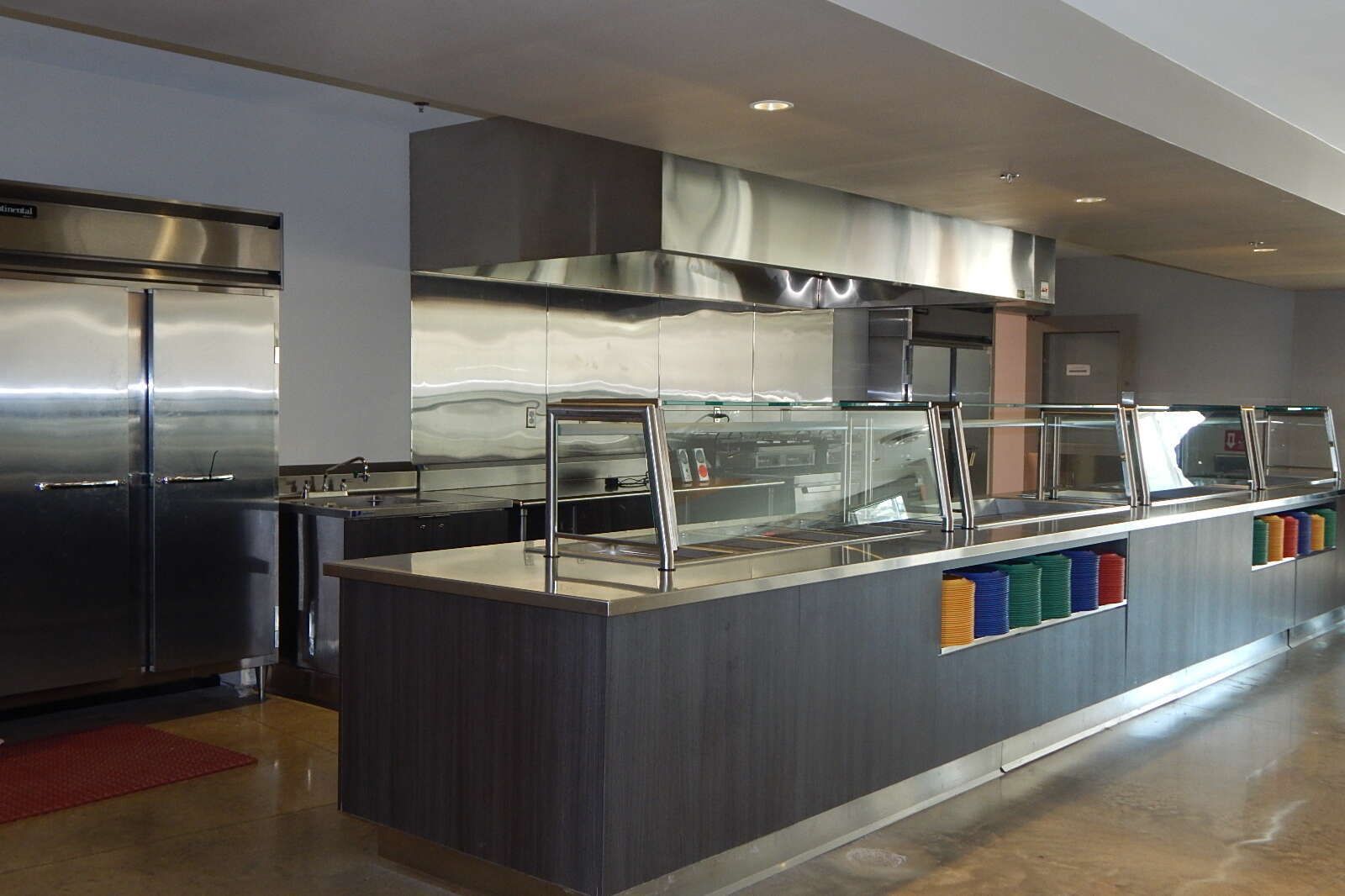 A modern, commercial cafeteria serving station with stainless steel appliances, glass guards, and a dark wood counter.