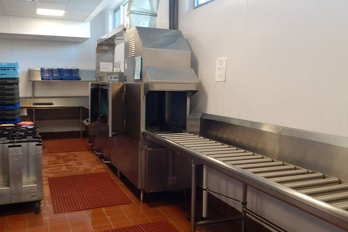 A commercial stainless steel dishwashing machine in a kitchen, featuring a metal conveyor belt and red floor mats.