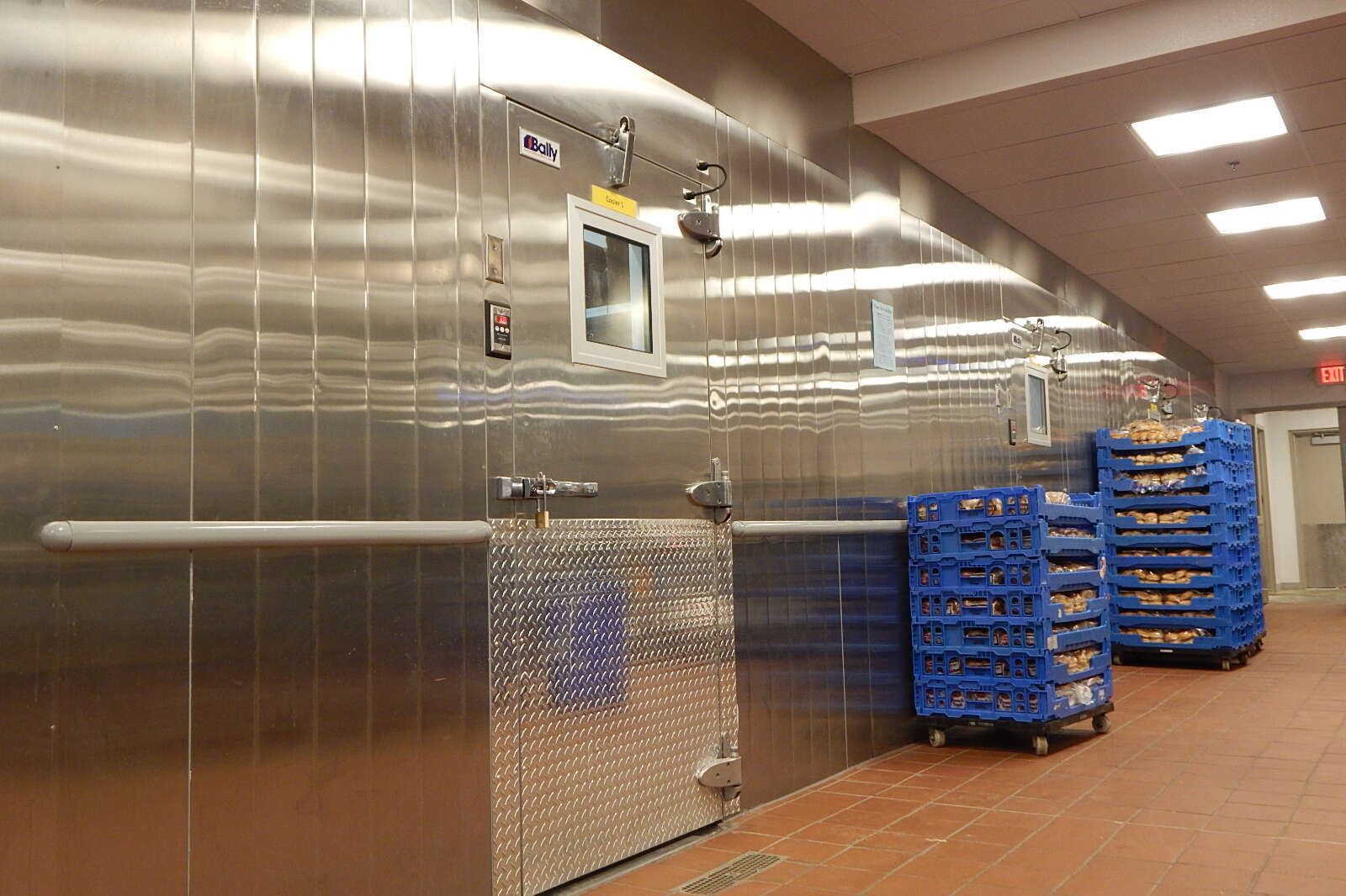 A commercial kitchen with stainless steel walls and a large walk-in cooler door beside stacks of blue plastic crates.