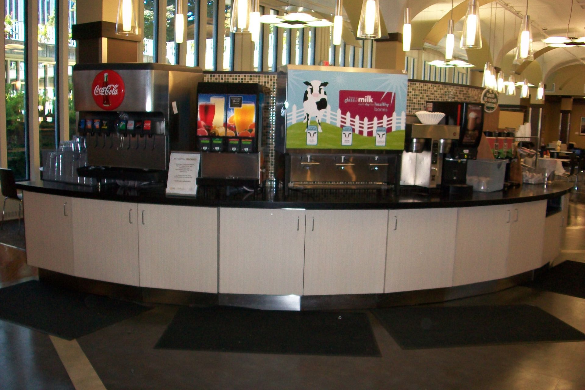 A curved beverage station in a dining hall featuring Coca-Cola and milk dispensers on a white counter.