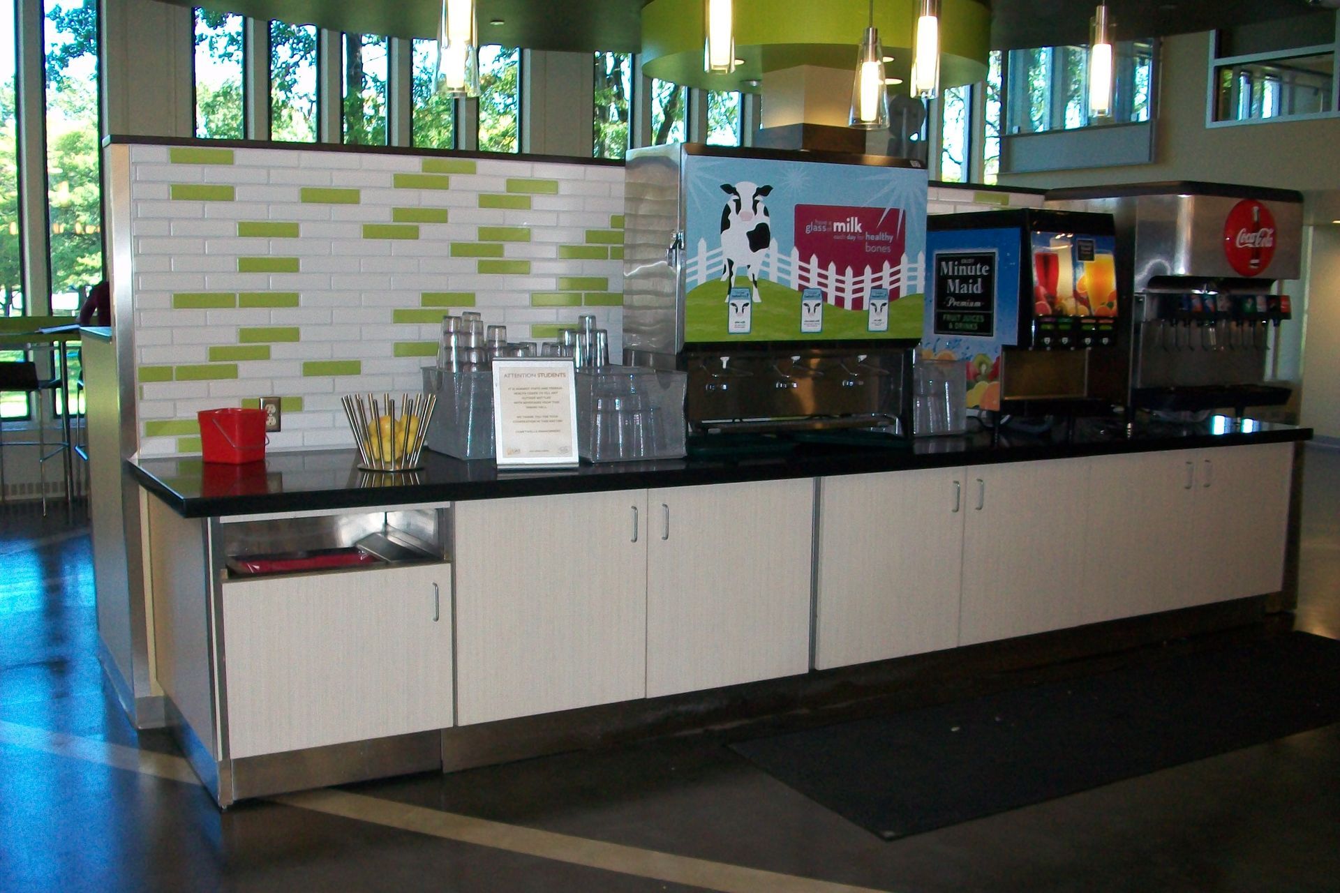 A drink station with milk and soda dispensers on a black counter, set against a tiled wall in a brightly lit dining area.
