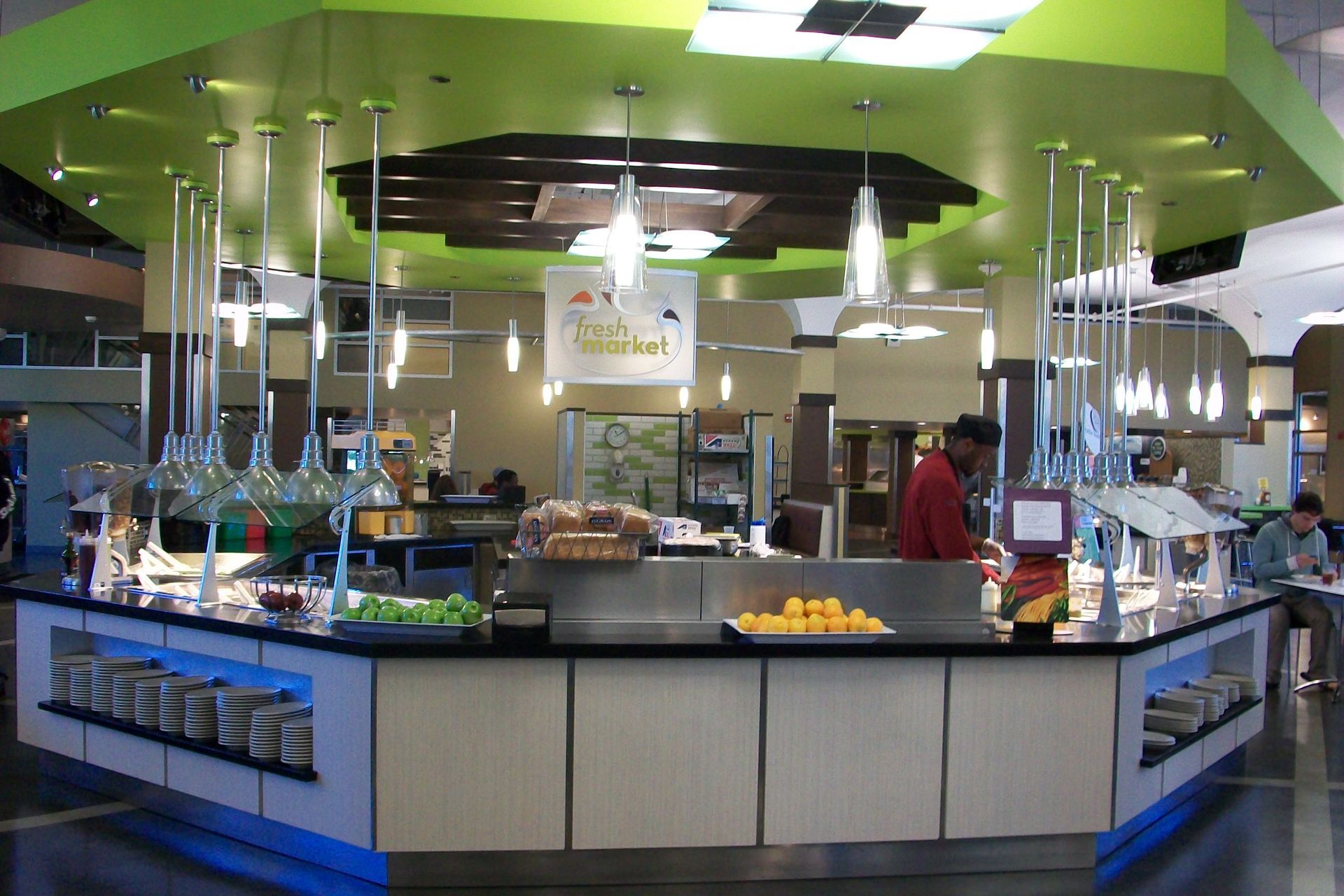 A modern salad bar island with green lighting, pendant lamps, and fresh produce inside a brightly lit dining area.