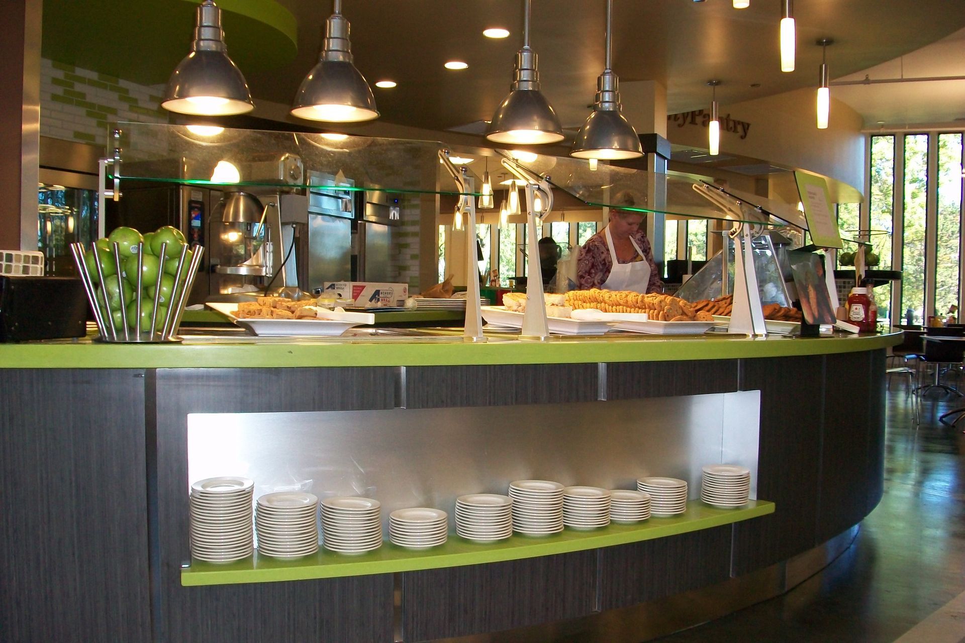 A buffet counter with a green surface, a basket of limes, and stacks of plates on a lower shelf in a modern dining hall.