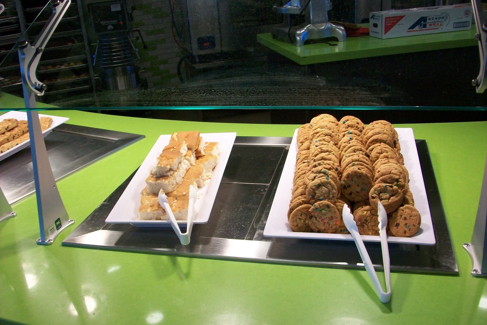 Two platters of cookies and treats on a bright green cafeteria counter with white serving tongs.
