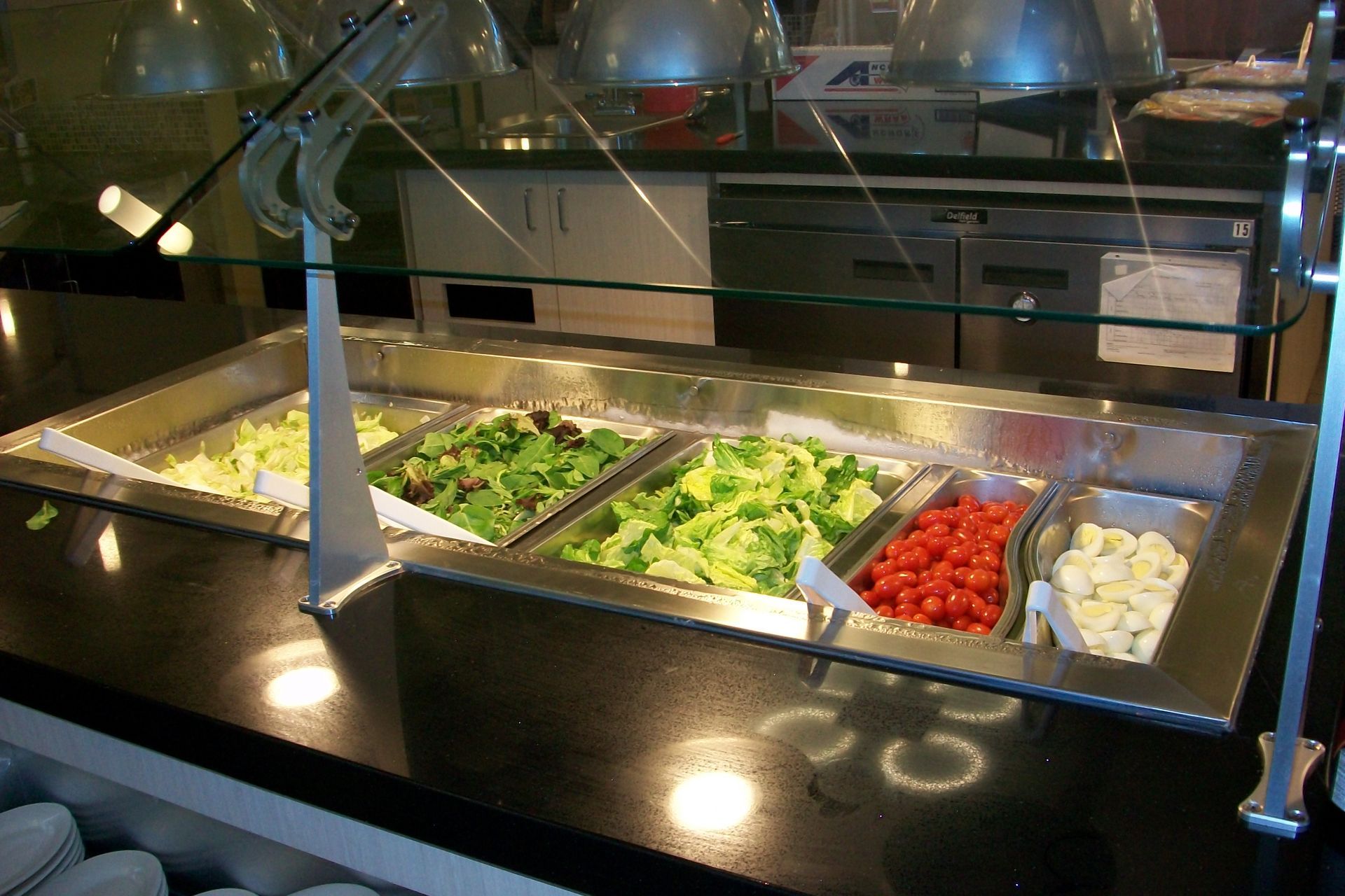 A salad bar with containers of mixed greens, lettuce, cherry tomatoes, and sliced mushrooms under glass shields.