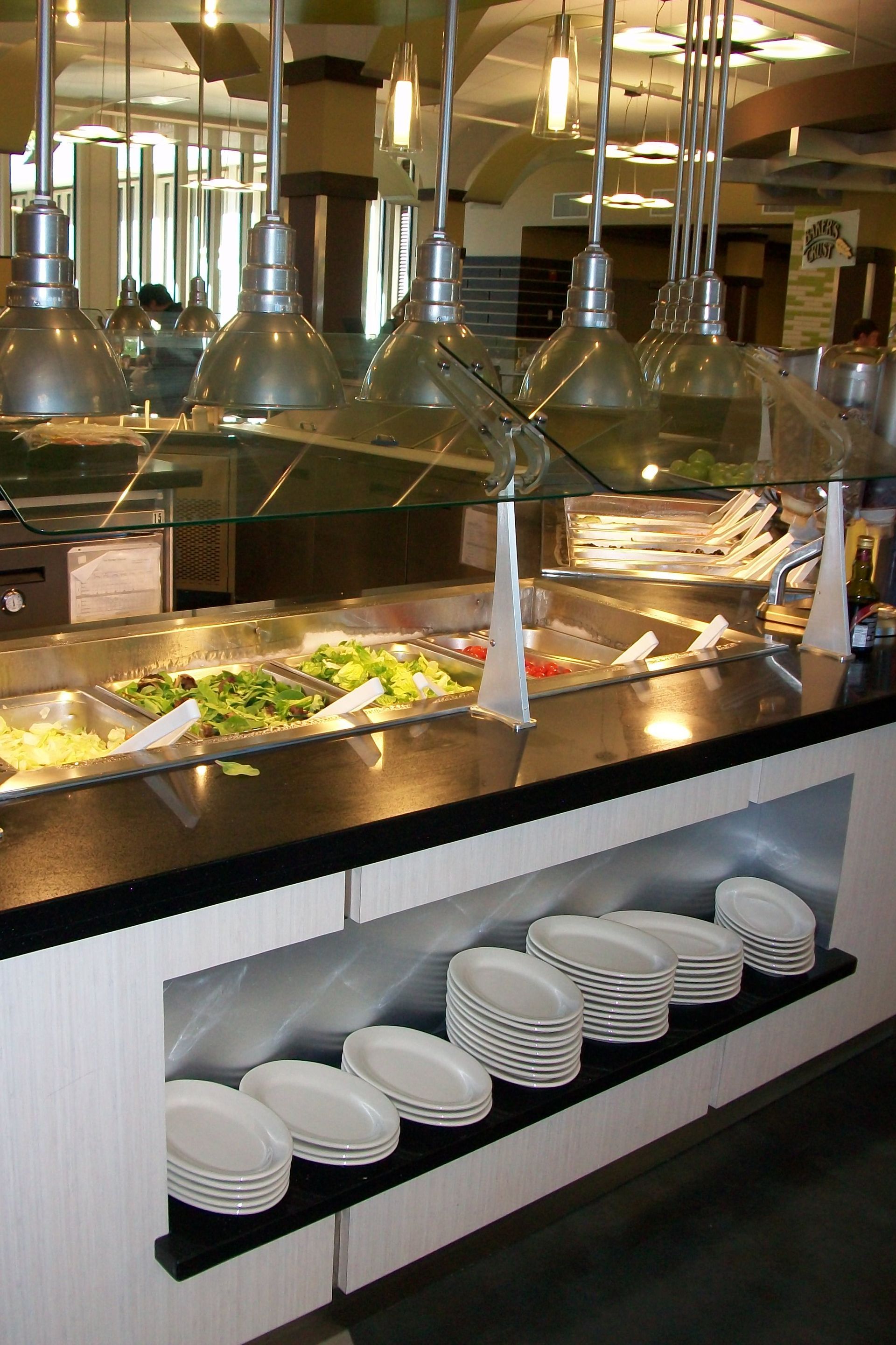A buffet salad bar with overhead lights, metal containers of food, and stacks of white plates on a lower shelf.