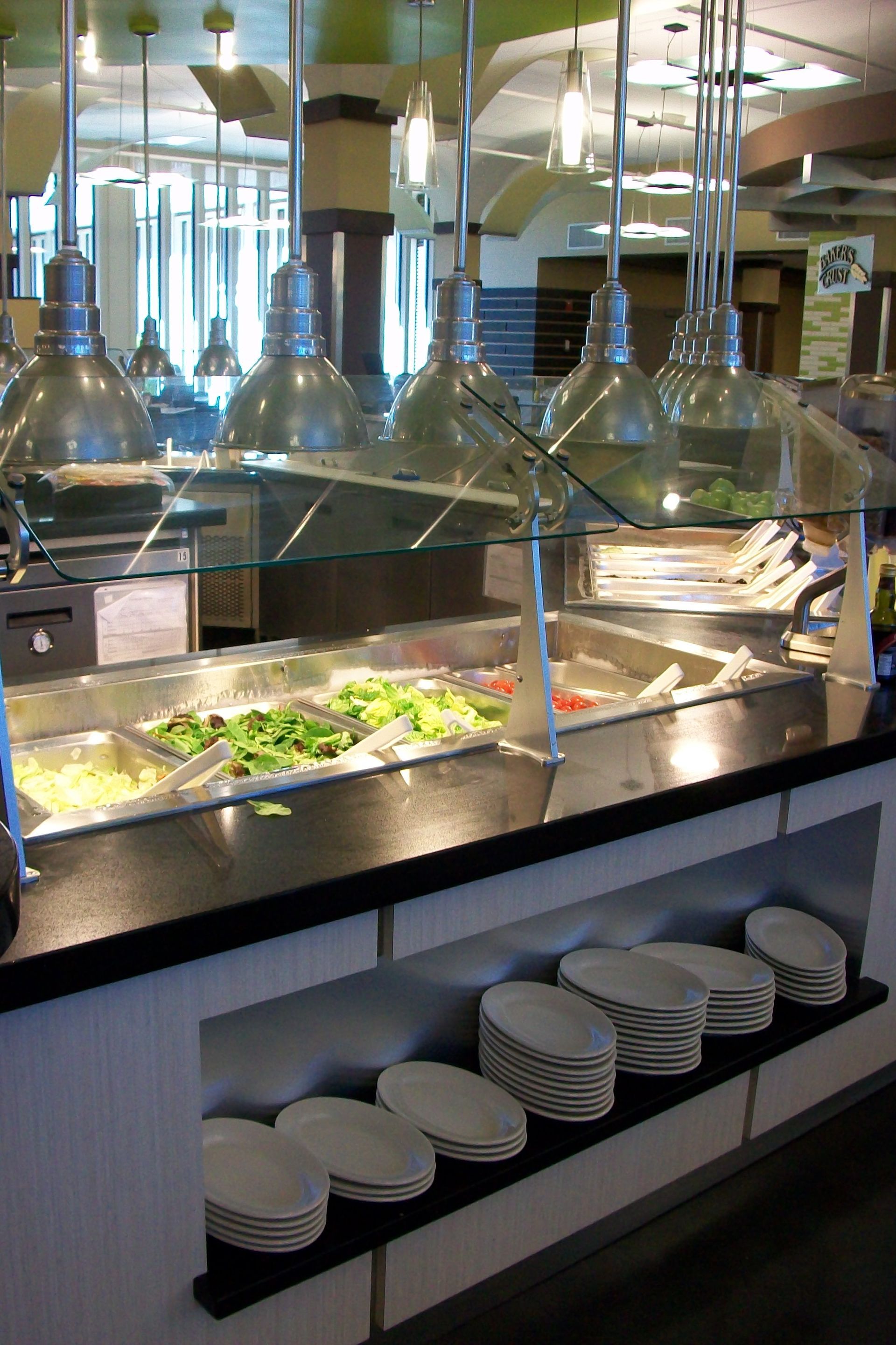 A salad bar with hanging metal pendant lights and stacks of white plates on a shelf below.