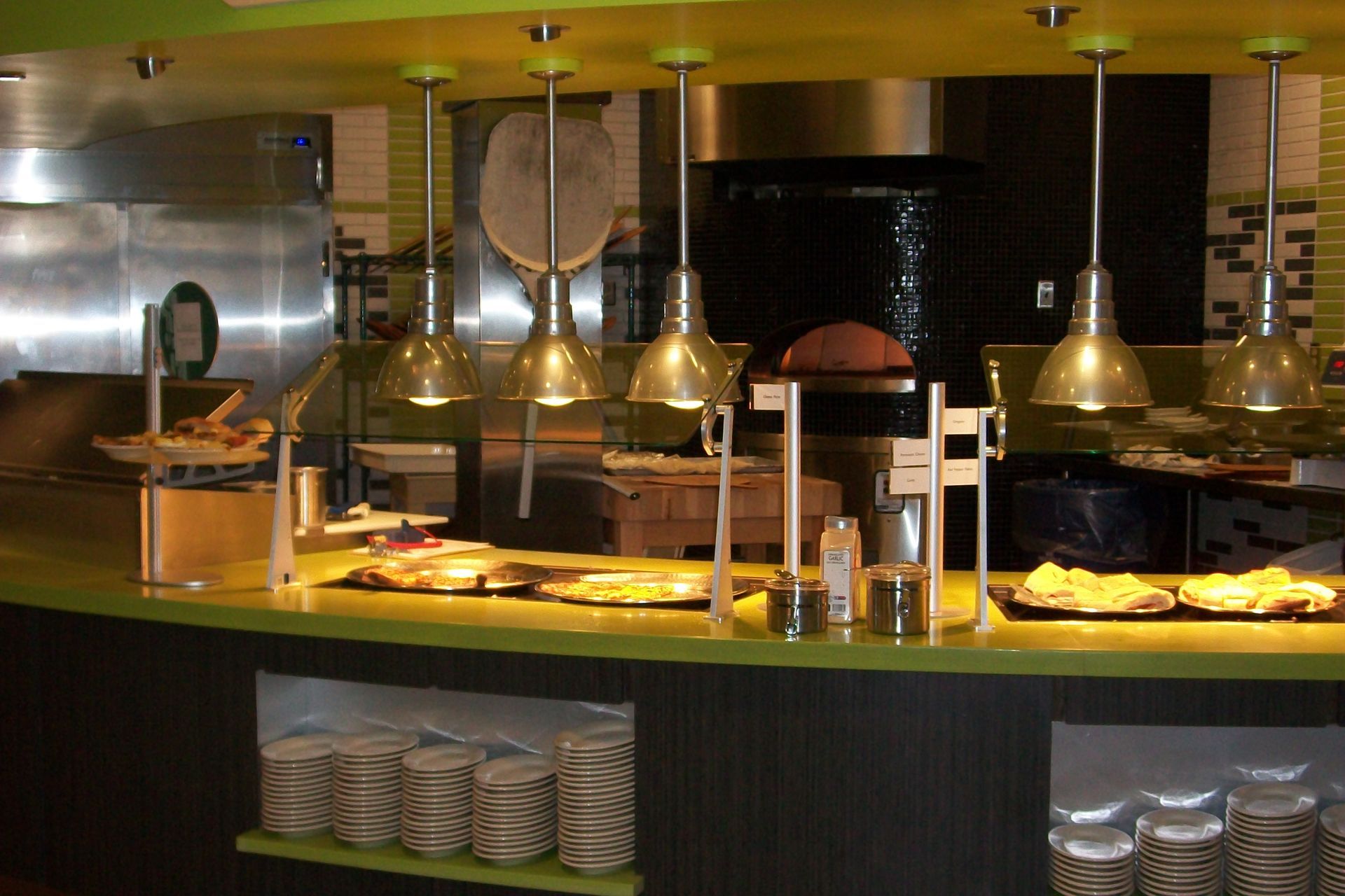 A buffet counter with warm hanging lights, stainless steel equipment, and stacks of plates on lower shelves.