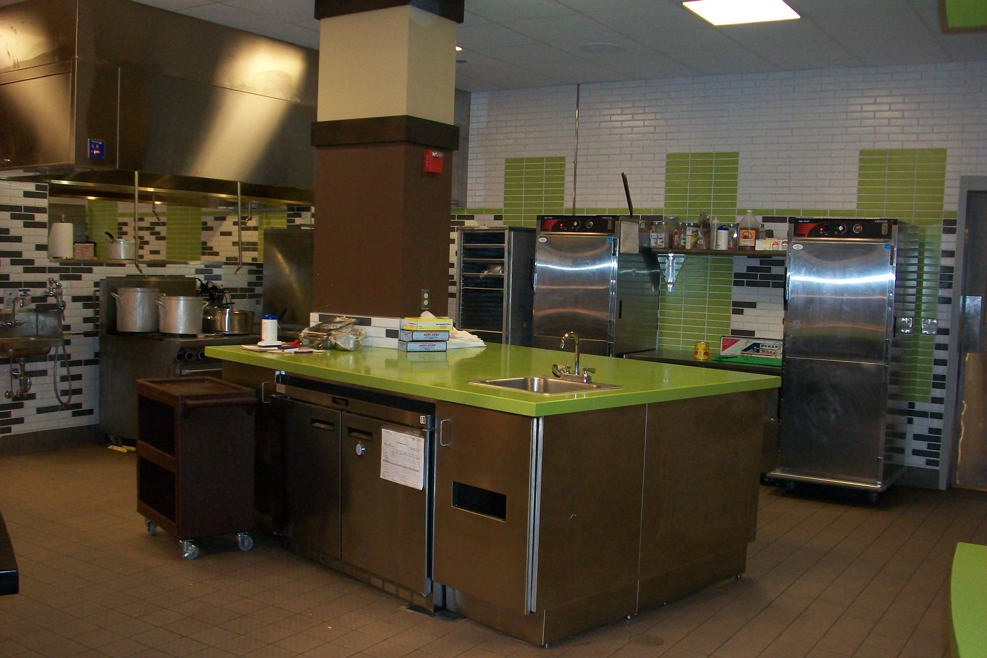 Commercial kitchen featuring stainless steel appliances, a green countertop island, and white and green tiled walls.