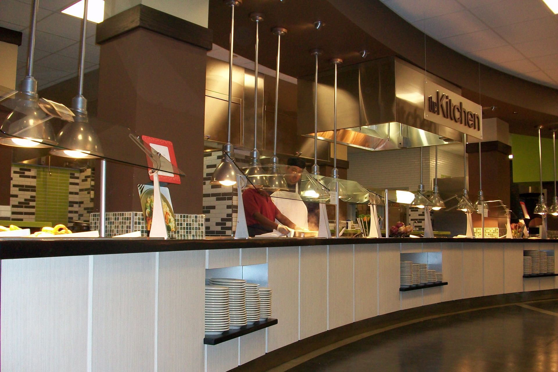 A service counter at a buffet-style restaurant with hanging pendant lights, food warmers, and a staff member cooking.