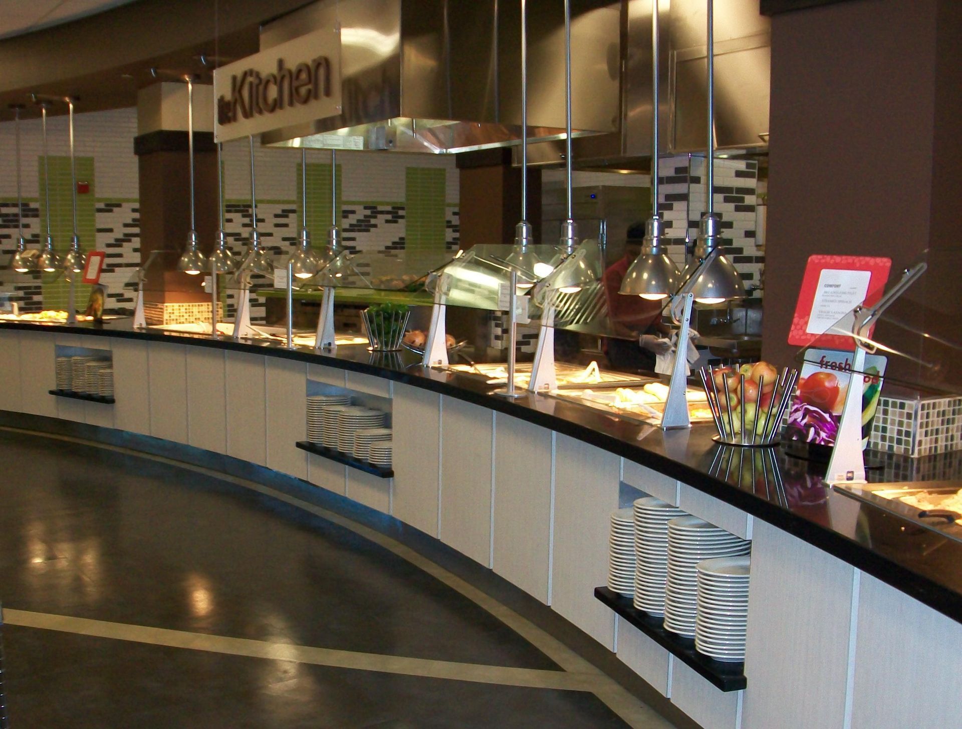 A curved buffet service counter in a modern dining area, featuring hanging heat lamps and stacked white plates.