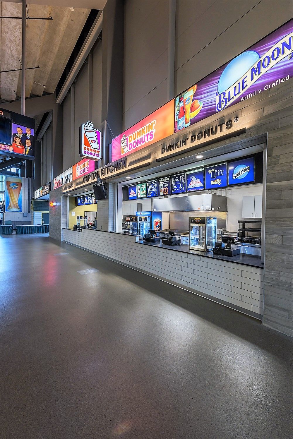 An indoor concession stand with illuminated signs for Blue Moon and Dunkin’ along a long counter in a stadium concourse.