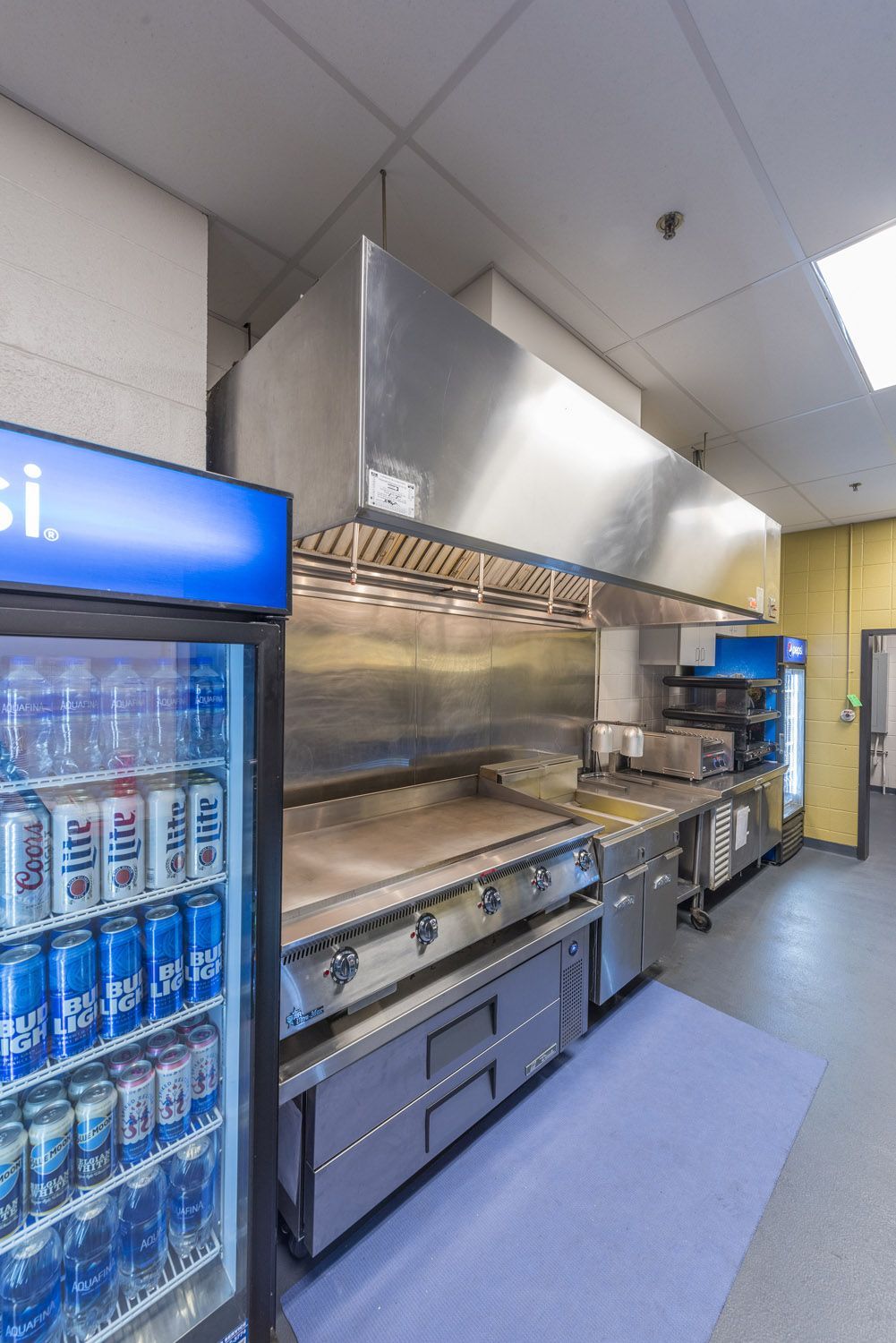 A commercial kitchen space featuring a metal exhaust hood over a griddle, beside a refrigerated drink display case.