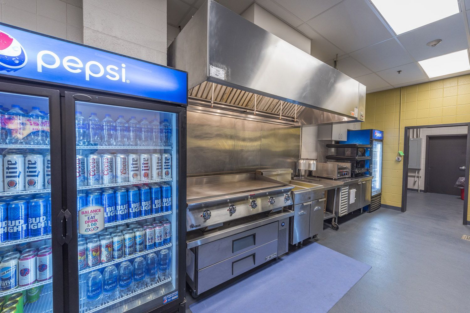 A commercial kitchen with a stainless steel range and hood, next to a Pepsi-branded glass display refrigerator.