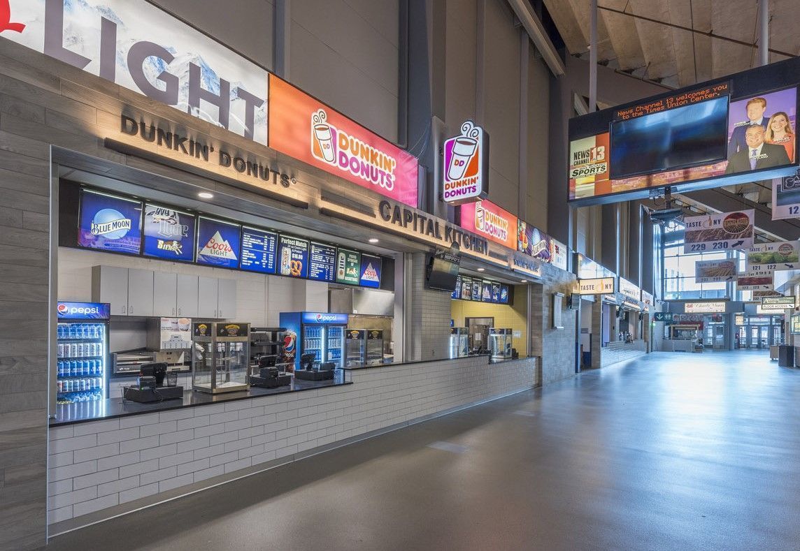 A view of a Dunkin' Donuts concession stand inside a stadium concourse with digital menu boards and advertisements.