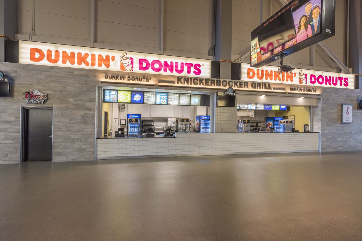 A Dunkin' Donuts concession stand in an indoor stadium or arena, featuring menu boards above a service counter.