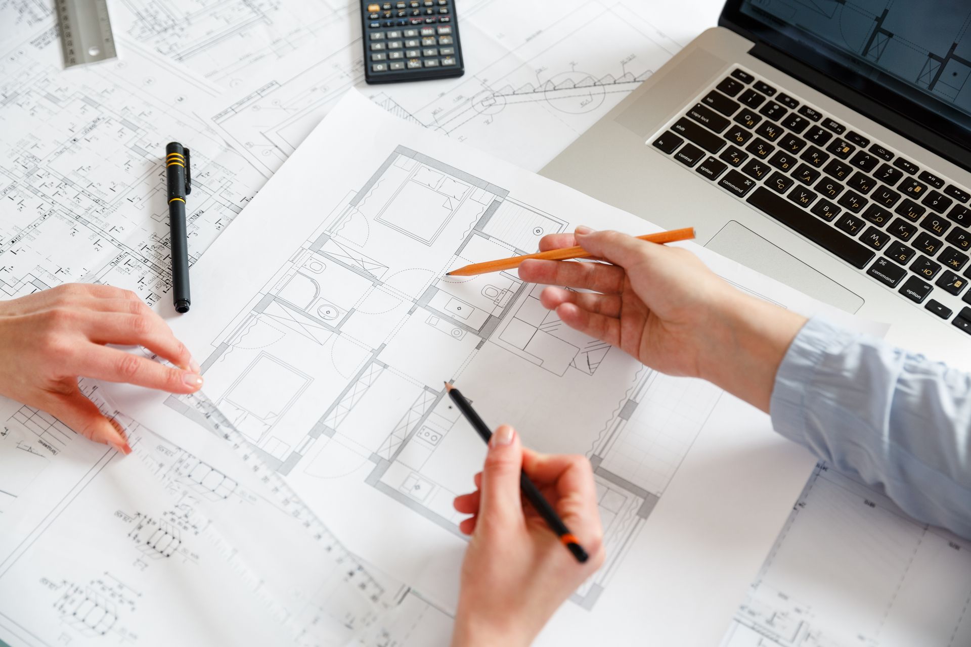 Two people reviewing architectural floor plans on a desk with a laptop, calculator, and drawing tools.