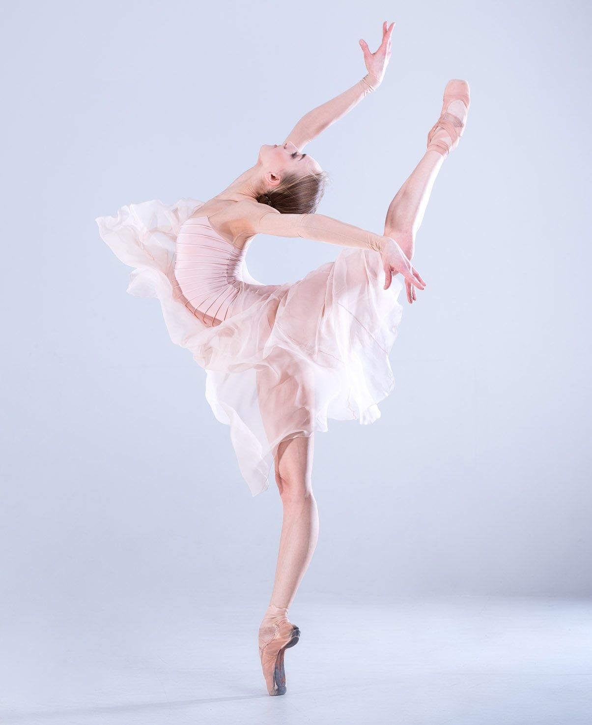 Ballerina in pink dress, balances en pointe with leg extended high; arms gracefully arched.