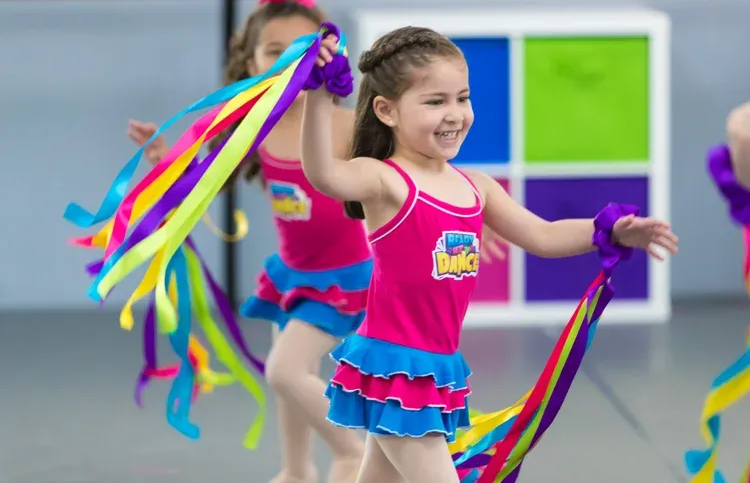 Two young girls in pink leotards dance with colorful ribbons, smiling. A dance studio backdrop is in view.