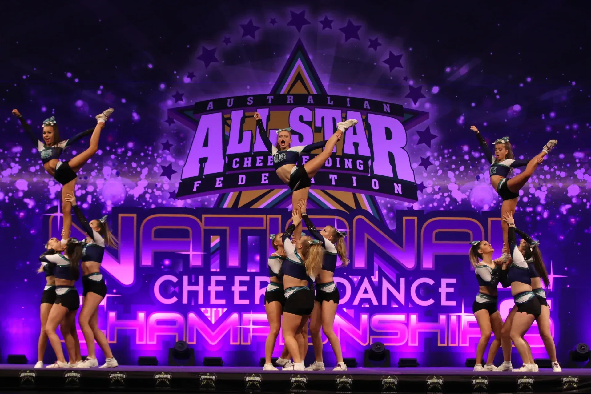 Cheerleaders perform at a national championship, with a backdrop. They are in a pyramid formation, wearing black and blue.