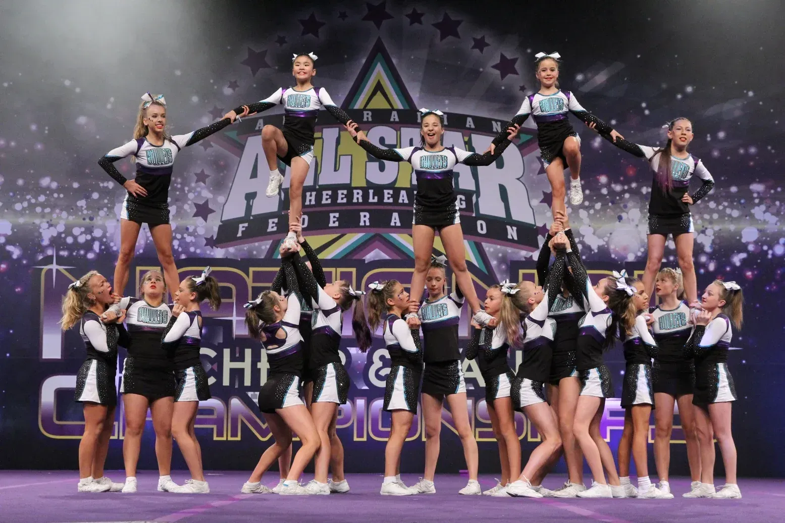 Cheerleaders in black and white uniforms performing a pyramid stunt on a stage at a competition.