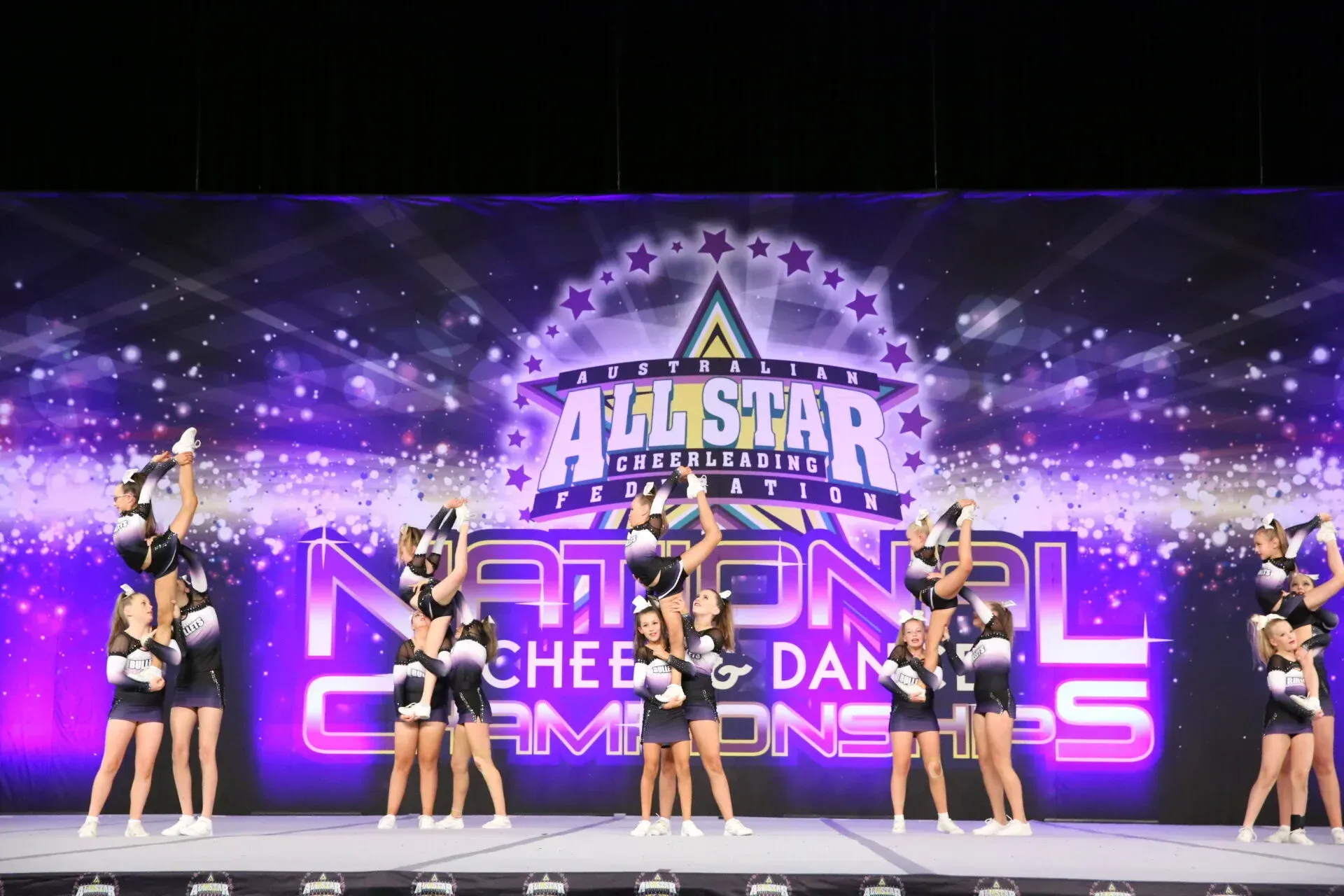 Cheerleading team performs stunts at a competition, with purple and black uniforms and a stage backdrop.