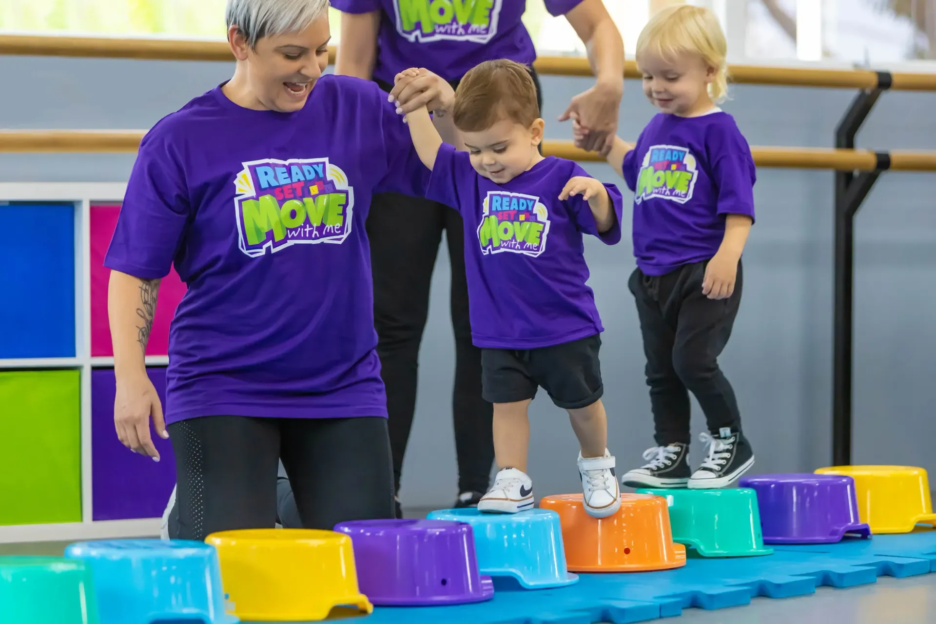 Woman helps children step on colorful blocks; dance studio.