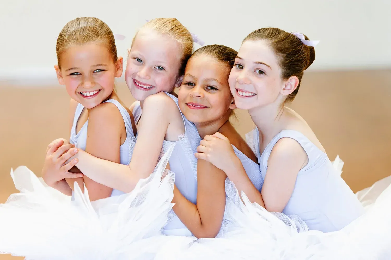Four young ballet students in white costumes embracing and smiling at Mathis Dance Studios