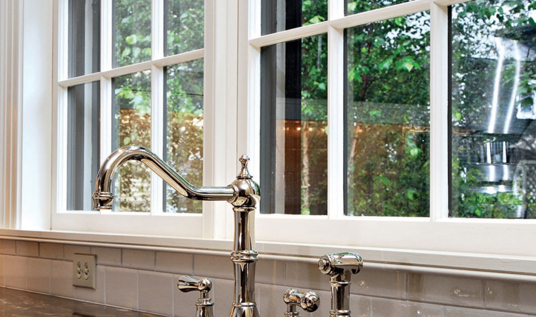Kitchen sink with chrome faucet in front of a white framed window with a view of greenery.
