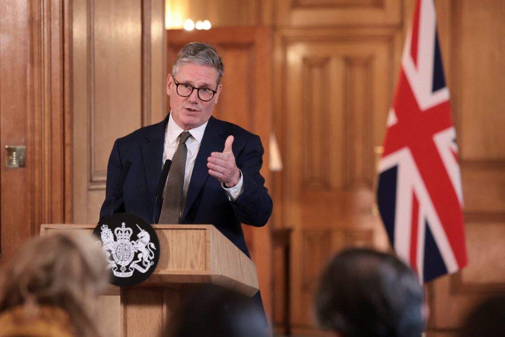 Man speaking at a podium with UK flag behind him in a wood-paneled room
