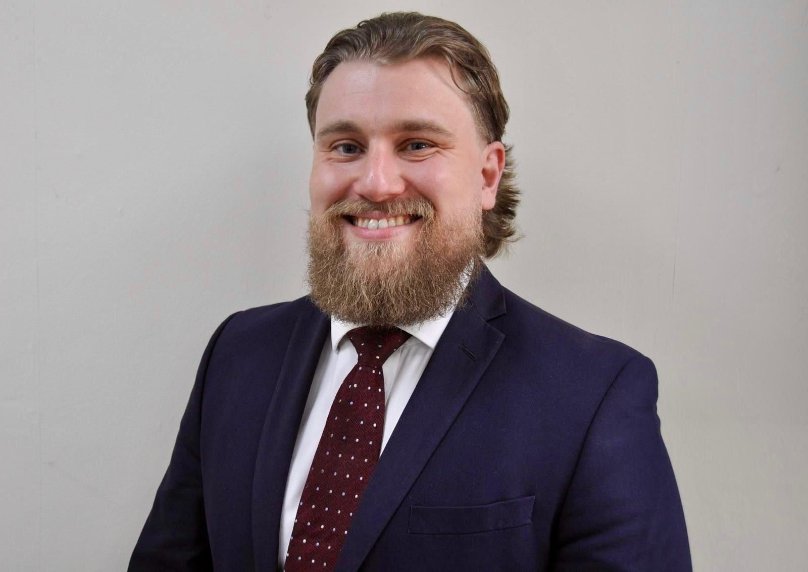 A smiling person with a beard and tied-back hair, wearing a dark blue suit, white shirt, and patterned burgundy tie.
