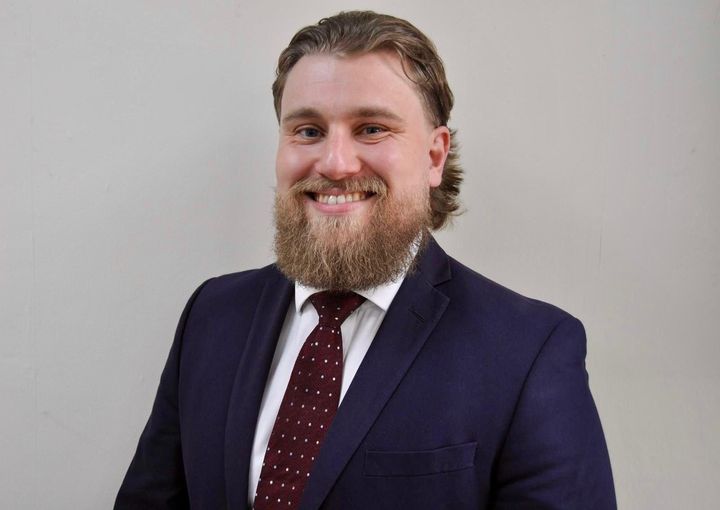 A smiling person with a beard and tied-back hair, wearing a dark blue suit, white shirt, and patterned burgundy tie.
