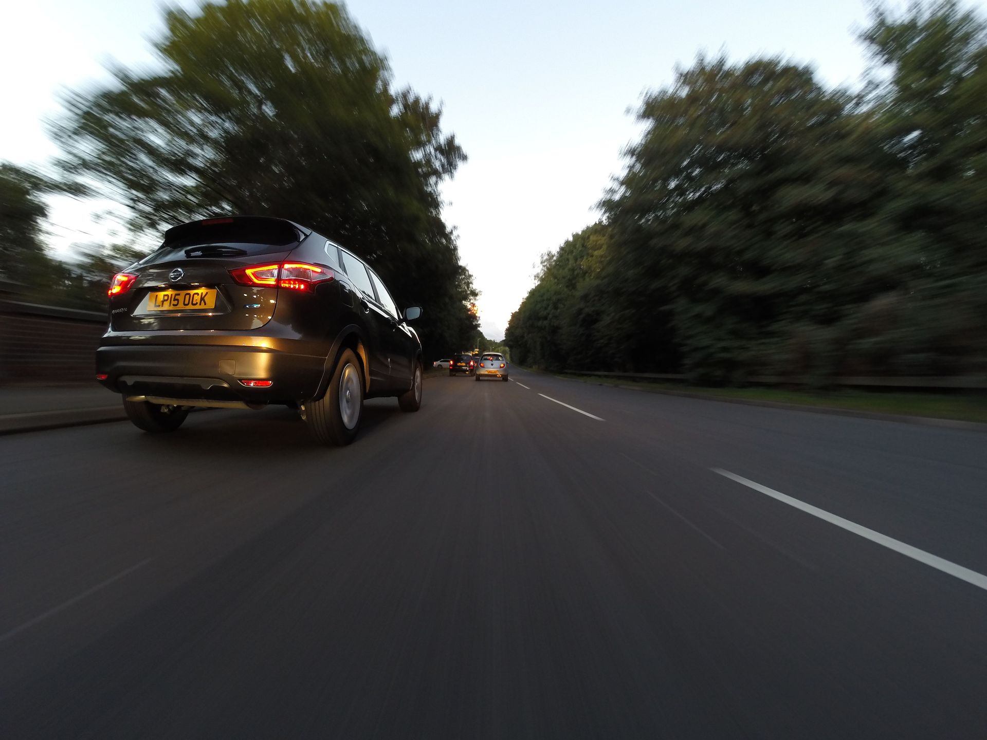 Black SUV driving on a tree-lined road at dusk, viewed from behind.