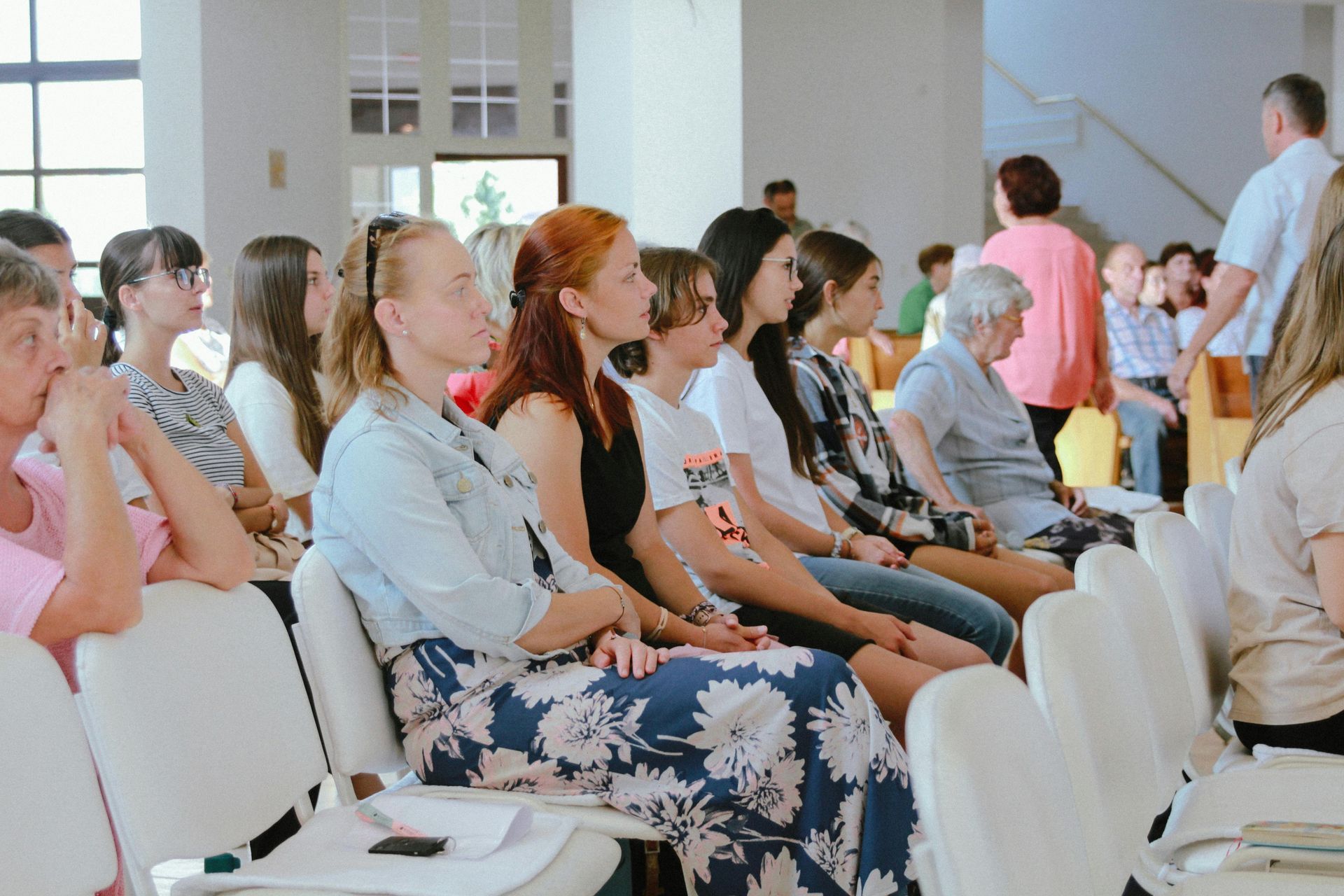 People seated in rows of white chairs at an indoor event, listening attentively.