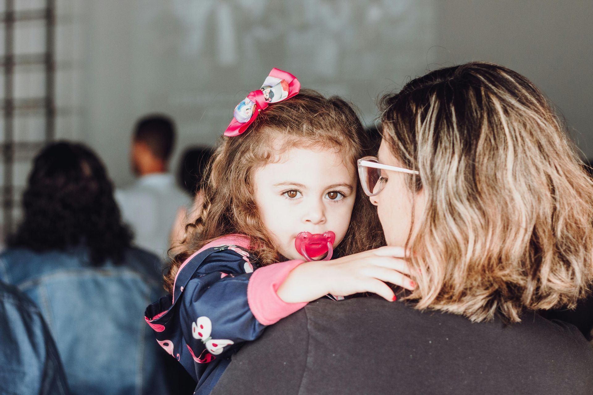 Child with pink bow and pacifier resting on an adult’s shoulder in a softly blurred indoor setting