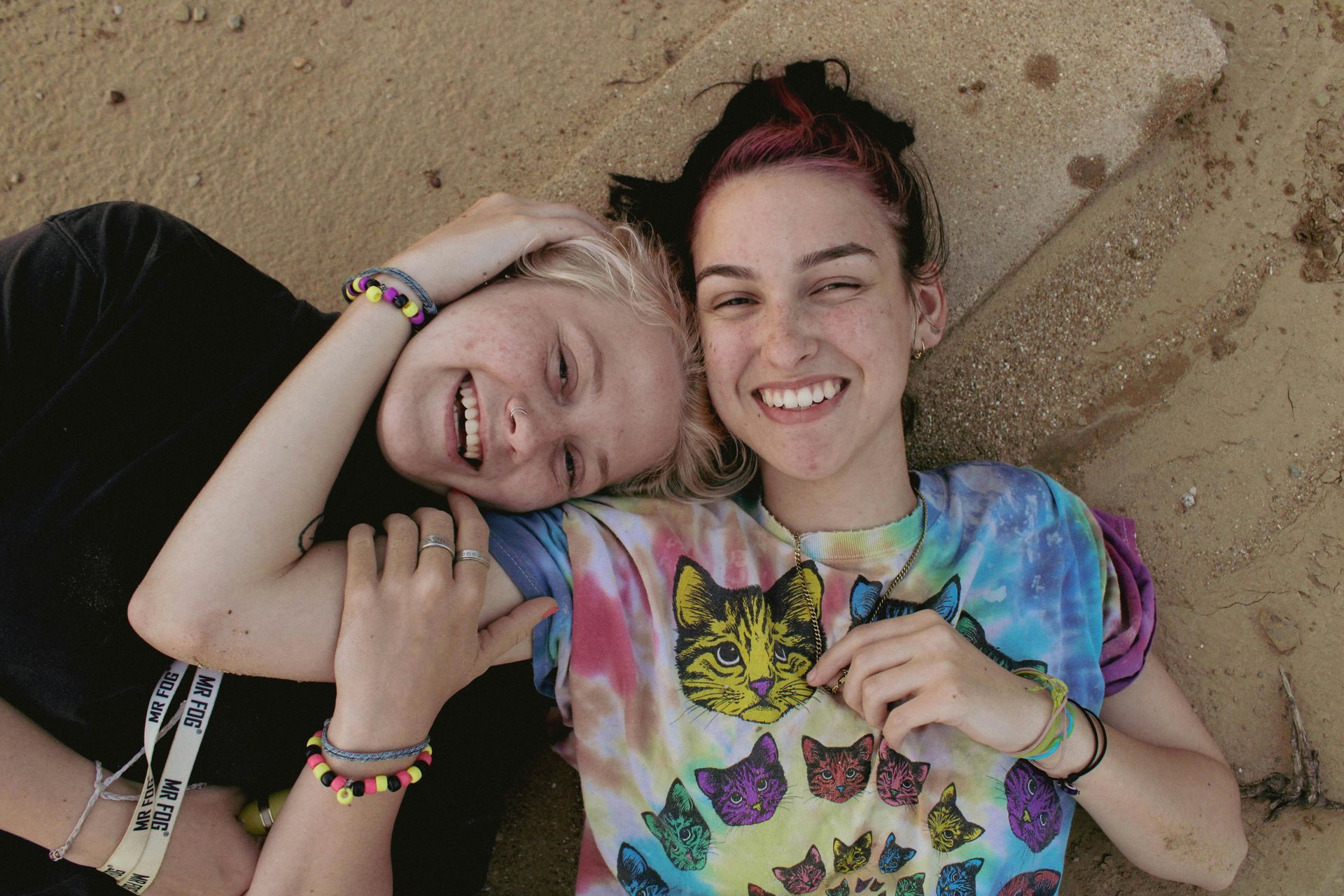 Two smiling friends lying on sand, one in a tie-dye shirt, looking at the camera.