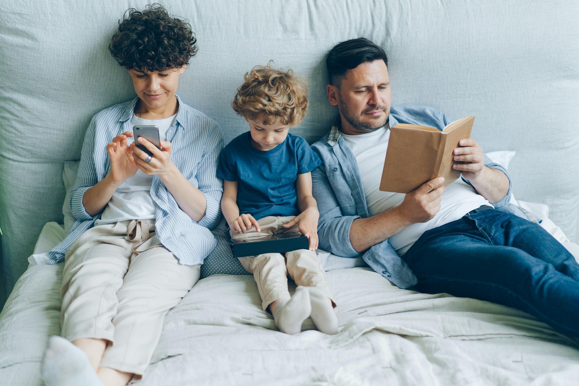 Three family members sitting on a bed, two using phones and one reading a book.