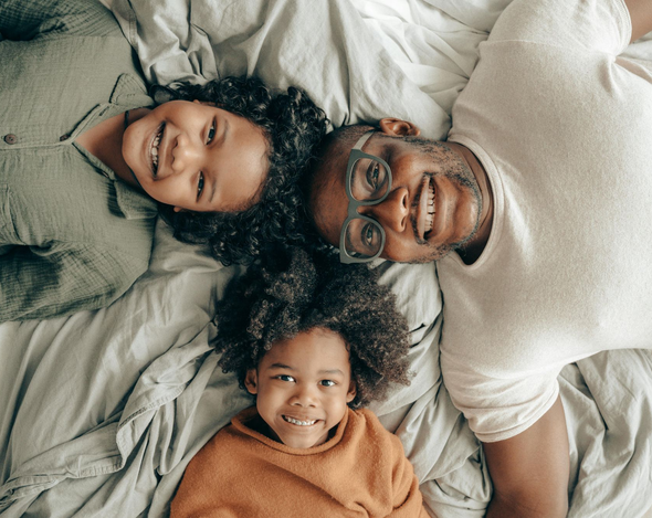 Three smiling people lying on a bed, heads together on white sheets.