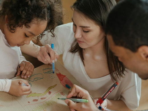 Family gathered around a table, drawing and painting on a large sheet of paper with markers and brushes.
