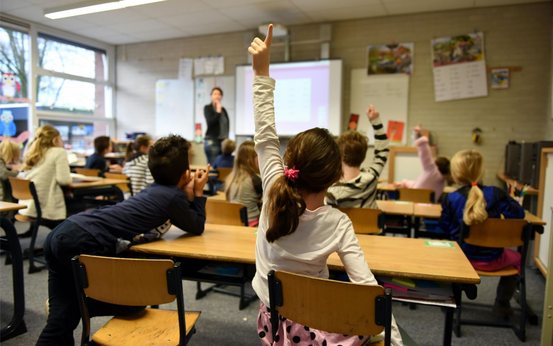 Classroom with students seated at desks, one child raising a hand near a whiteboard and projection screen.