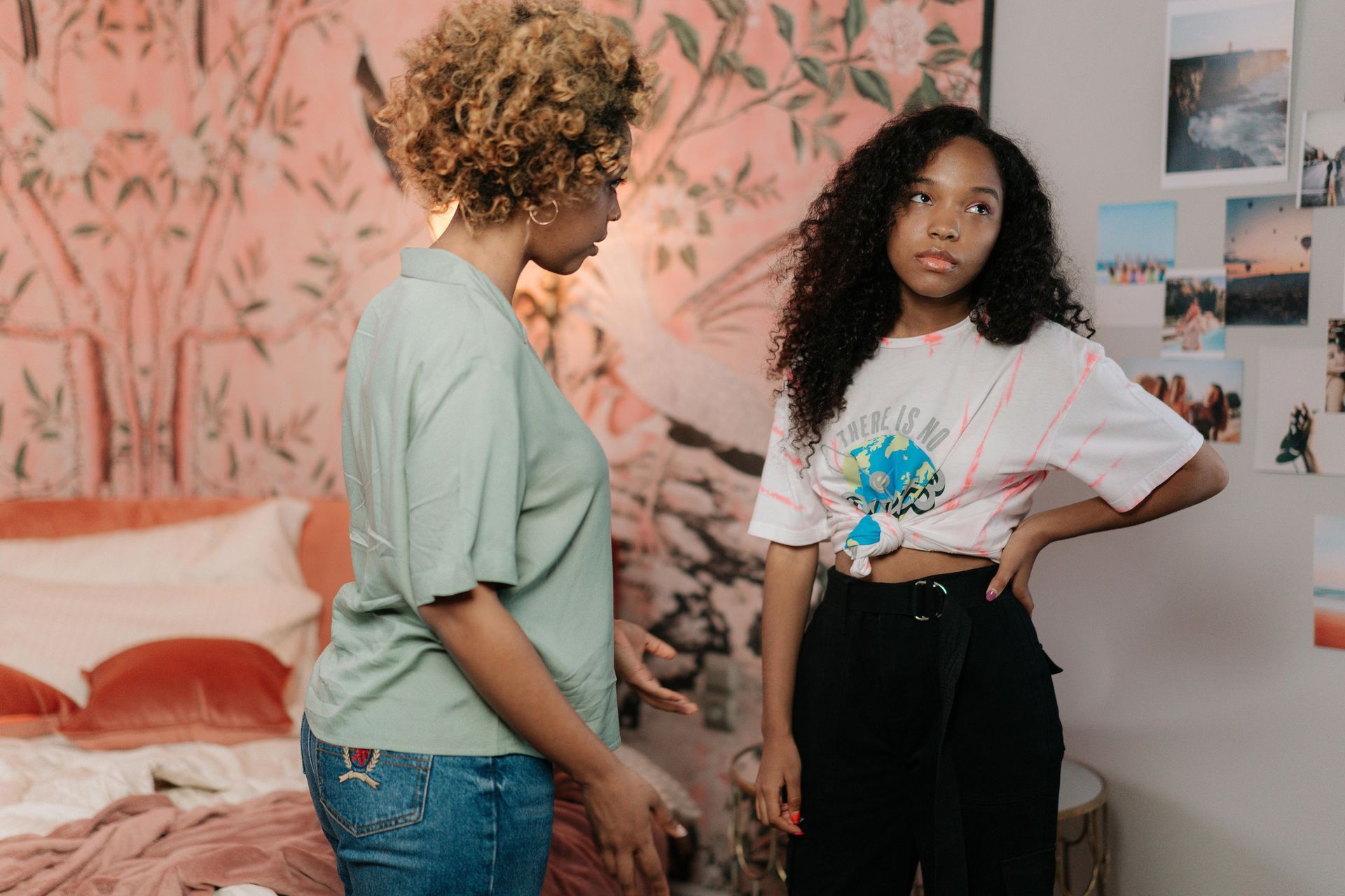 A parent and daughter standing in a bedroom, one in a green shirt facing a woman in a white graphic tee.