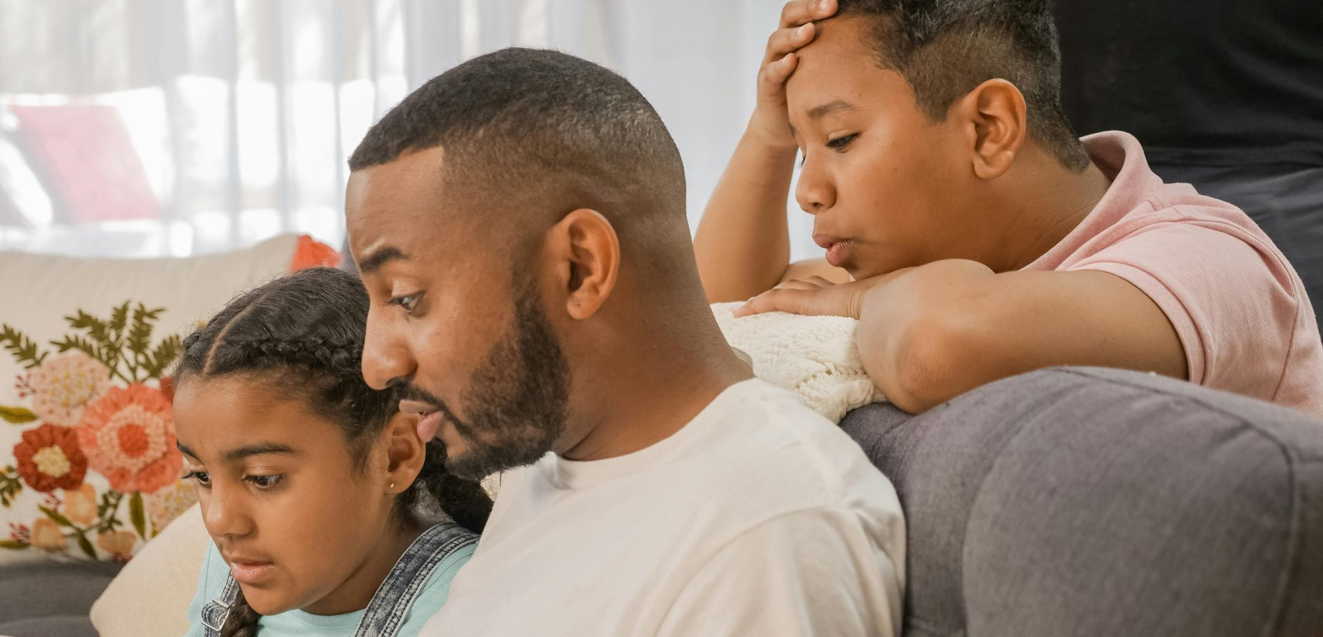 A father with his son and daughter sitting closely on a couch, looking at a device with serious expressions.