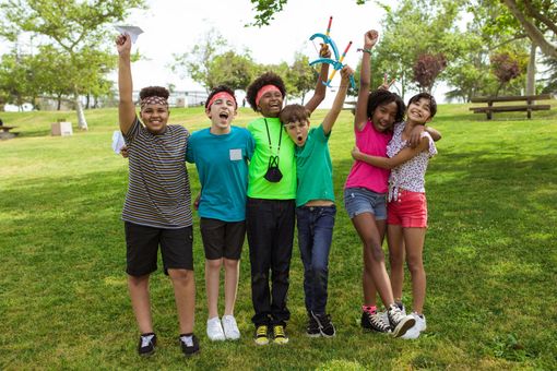 Six friends smiling and cheering together in a sunny park, standing on grass with trees behind them.