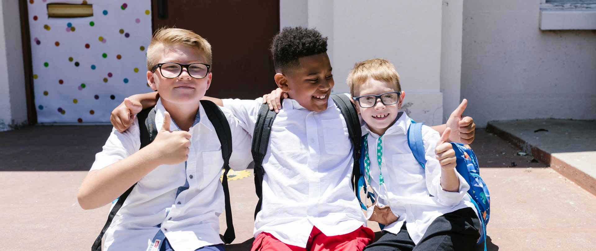Three smiling school friends sitting together outdoors, giving thumbs up, with backpacks on.