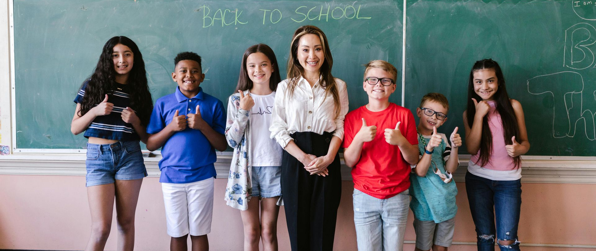 Six schoolchildren stand smiling in front of a chalkboard in a classroom with their teacher