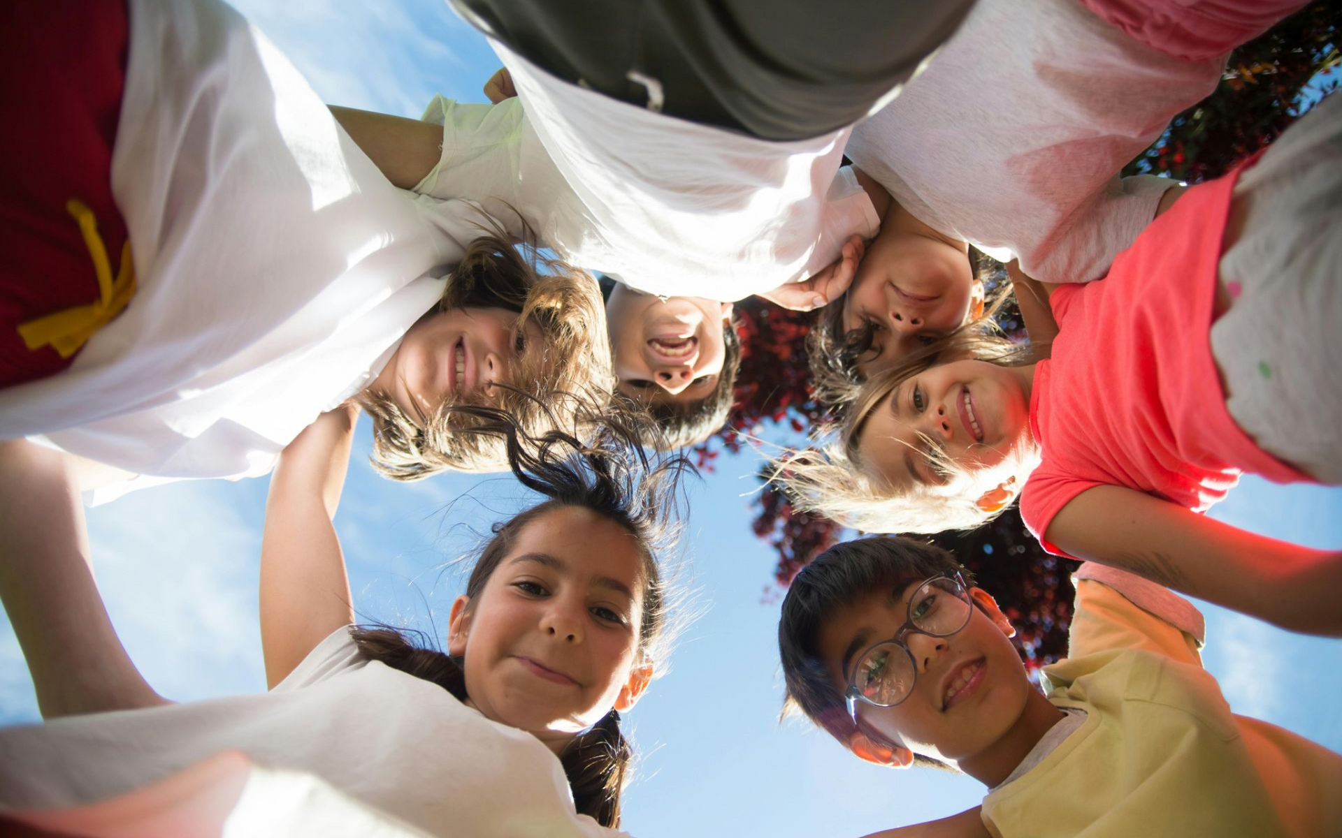 Group of smiling friends huddled in a circle, looking down at the camera against a blue sky