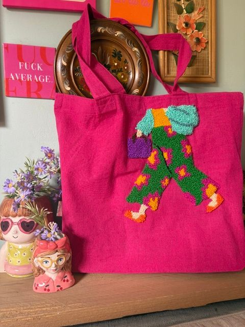 Een oranje draagtas met geborduurde kaarsen hangt aan de muur naast ingelijste kunst en paddenstoelbeeldjes.