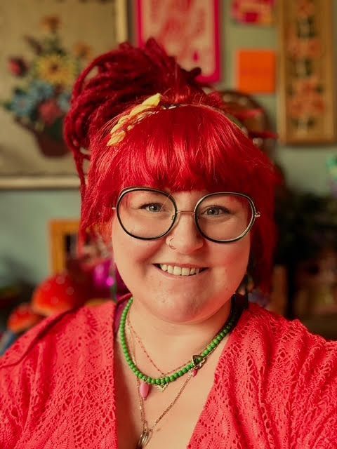 Een vrouw met rood haar en een bril glimlacht. Ze draagt een rode top en kralenkettingen, tegen een kleurrijke achtergrond.