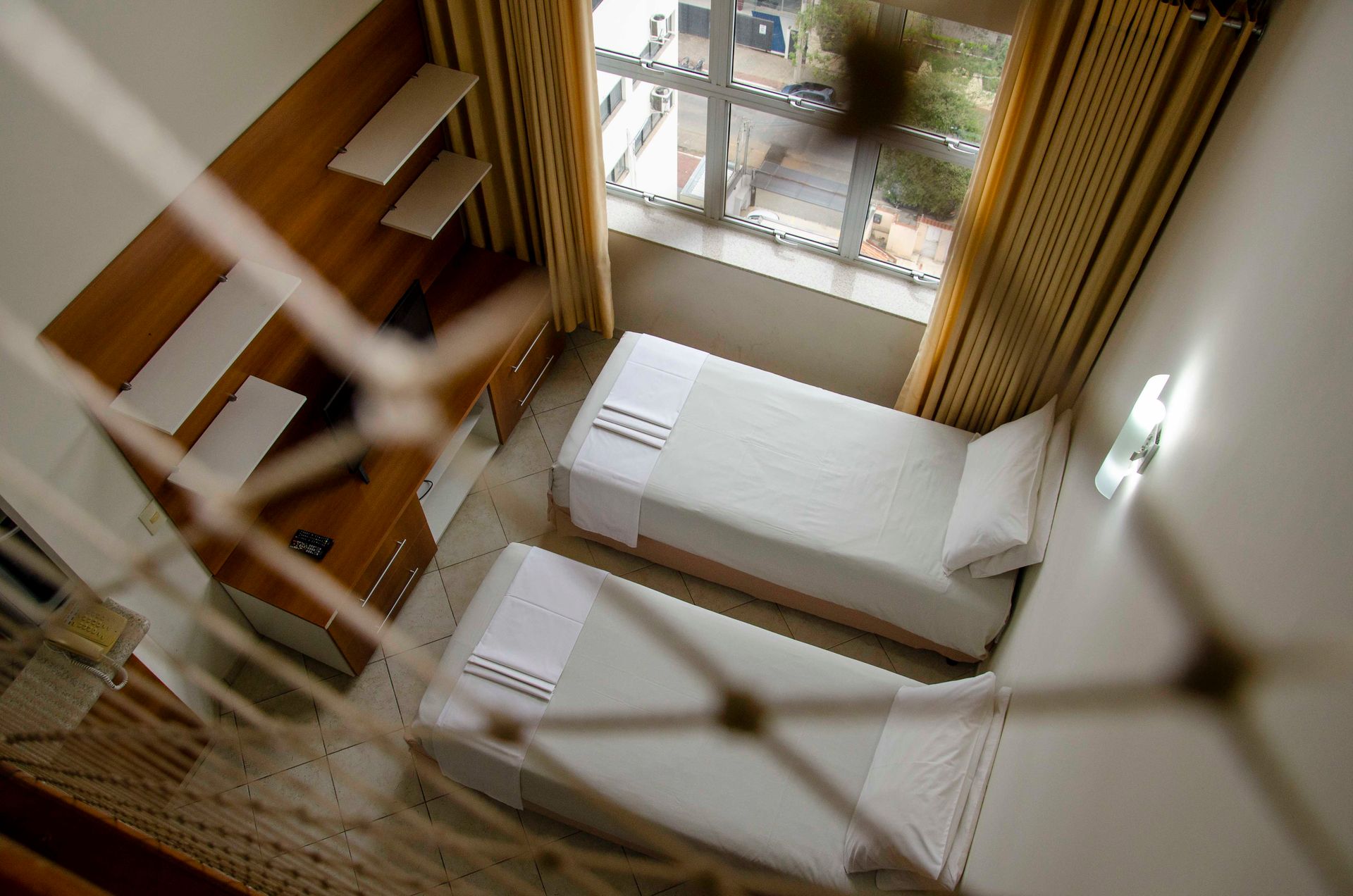 Quarto de hotel aconchegante com cama, área de estar, luminária e janela com cortina em tons suaves de bege.